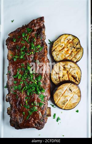 Vue de dessus de délicieuses côtes de porc grillées garnies de fruits frais persil servi avec des tranches d'aubergines grillées sur un plateau blanc Banque D'Images