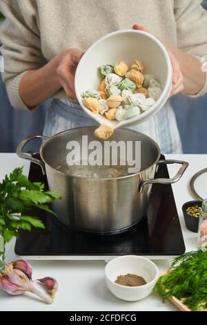 Femme sans visage en tablier versant des boulettes faites de la pâte avec des carottes et des épinards dans une casserole avec de l'eau bouillante sur la table souhaitent herbes fraîches et espèces Banque D'Images