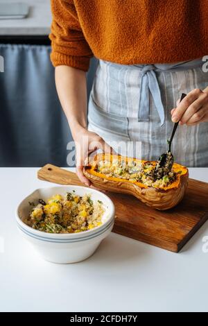 Crop femme de ménage remplissant le courge de noyer cendré cuit avec un mélange végétarien de quinoa et légumes à table blanche dans la cuisine maison Banque D'Images