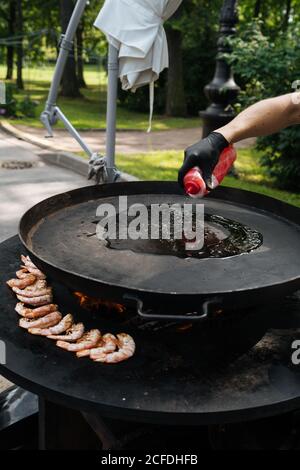 Du dessus du chef de la récolte en gant avec bouteille verser l'huile à la main sur la casserole près des crevettes sur le marché Banque D'Images