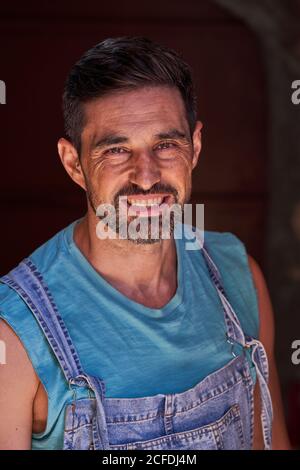 Joyeux barbu souriant ouvrier d'âge moyen portant une chemise bleue et combinaison de jeans avec fond sombre Banque D'Images