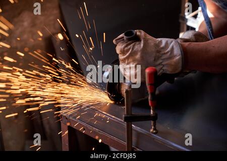 Coupe des mains du travailleur dans des lunettes de protection et des gants métal avec moulin à étincelles volantes pendant les travaux en atelier Banque D'Images