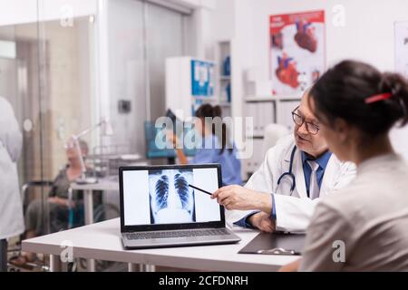 Un médecin mature pointant vers une tomodensitométrie des poumons du patient sur un ordinateur portable lors d'une visite médicale au bureau de l'hôpital. Medic en manteau blanc discutant avec une femme handicapée en fauteuil roulant et une infirmière en tenant des rayons X. Banque D'Images