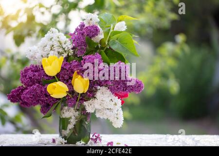 Printemps ou été. Un bouquet de lilas et de tulipes jaunes dans un pot en verre sur la table. Belles fleurs sur fond vert. Banque D'Images