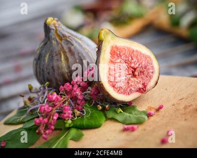 La moitié entière mûre et les figues en tranches parmi les feuilles vertes aromatisées d'herbes et de fleurs colorées sur planche à découper en bois contre mur blanc flou Banque D'Images