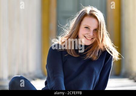 Portrait d'une jeune femme aux cheveux rougeâtres et aux taches de rousseur assis sur le sol en souriant à la caméra Banque D'Images