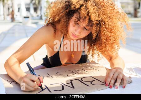 Femme ethnique sérieuse avec la coiffure afro créant carton poster avec inscription black lives matter assise pendant la démonstration Banque D'Images