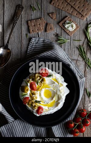 Vue de dessus des craquelins croquants et du romarin placés sur du bois table près de la serviette et assiette de yaourt de labneh avec tomates et olives Banque D'Images