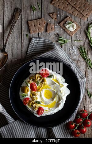 Vue de dessus des craquelins croquants et du romarin placés sur du bois table près de la serviette et assiette de yaourt de labneh avec tomates et olives Banque D'Images