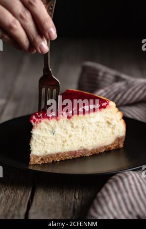 Croquer la femelle avec une fourchette en mangeant un délicieux cheesecake maison avec du rouge confiture de baies servie sur une assiette noire sur une table en bois Banque D'Images