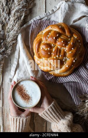 Par le dessus de la récolte femelle en chandail beige tenant le gobelet de cappuccino chaud tout en étant assis à une table en bois avec frais pain tressé fait maison placé sur le tissu à côté de la fourrure d'animal Banque D'Images
