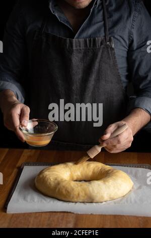 Petit chef cuisinier en tablier noir graissant du pain rond non cuit avec jaune d'œuf tout en étant debout sur une table en bois Banque D'Images