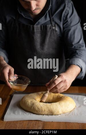 Petit chef cuisinier en tablier noir graissant du pain rond non cuit avec jaune d'œuf tout en étant debout sur une table en bois Banque D'Images