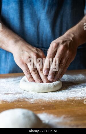 un homme méconnu en tablier aplatit la pâte molle sur la table avec farine tout en cuisant de la pizza Banque D'Images