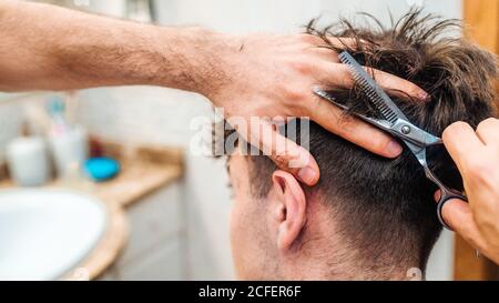 Vue arrière d'un homme méconnaissable en train de réaliser une coupe de cheveux à un type utilisant ciseaux contre l'intérieur flou de la salle de bains claire à la maison Banque D'Images