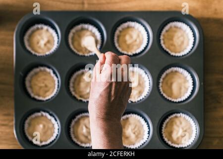 Vue de dessus de la femme anonyme de ménage âgée debout à la table avec moule à muffins et pâte à frire avec une cuillère dans des sous-moules à petits gâteaux Banque D'Images