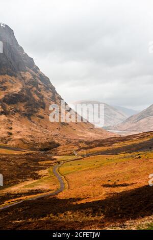 Route étroite et sinueuse traversant un terrain vallonné avec de l'herbe sèche Parmi les montagnes rocheuses en journée de printemps nuageux dans les Highlands écossais Banque D'Images