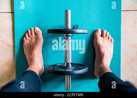 De ci-dessus crop anonyme appâted athlète masculin dans sportswear debout sur un tapis sport bleu vif contre une haltère pliable noire pendant préparation à l'entraînement à la maison Banque D'Images