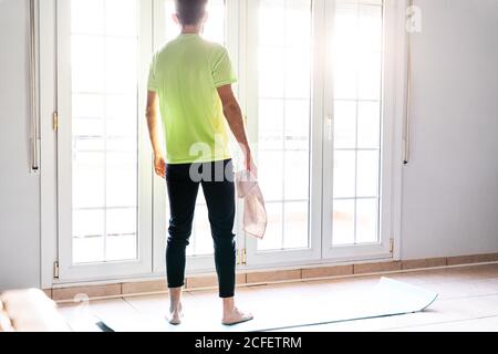 Vue arrière de la récolte pieds nus sportif en tenue active avec serviette donnant sur la fenêtre avec intérêt et réflexion pendant le repos après un entraînement difficile à la maison Banque D'Images