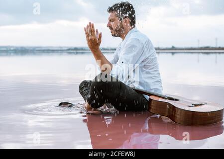 homme en chemise blanche et bretelles jetant de l'eau sur son visage tout en étant assis sur la plage avec de l'eau rose près de la guitare acoustique entouré d'un paysage de nuages majestueux et doux Banque D'Images