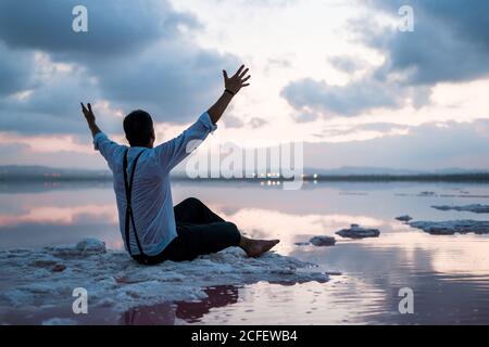 De derrière l'homme dans les vêtements mouillés regarder et s'asseoir avec les mains débordues au large au coucher du soleil nuageux Banque D'Images