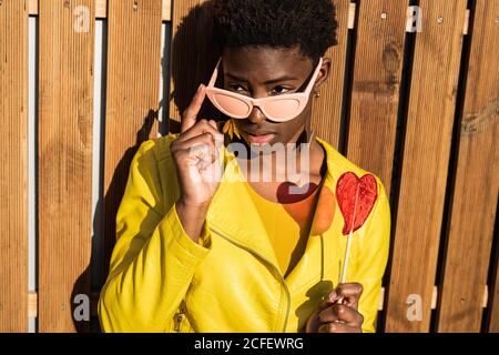 Femme afro-américaine tendance en lunettes de soleil dans la veste jaune appréciant coeur en forme de Lollipop par clôture en bois Banque D'Images