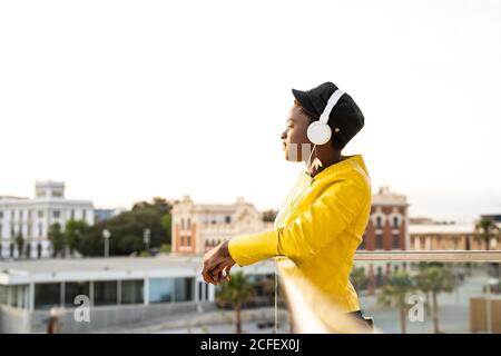 Vue latérale femme afro-américaine dans une veste tendance Ecoute de la musique dans un casque tout en s'inclinant sur un balcon en verre et en regardant loin sur un arrière-plan flou Banque D'Images