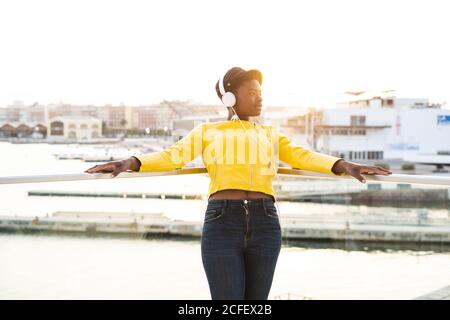 Contenu Afro-américaine femme dans une veste tendance écouter de la musique dans un casque tout en s'inclinant sur un balcon en verre et en regardant loin sur un arrière-plan flou Banque D'Images