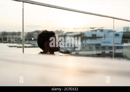 Vue latérale sélective d'une femme afro-américaine sereine qui se rafraîchissant sur un balcon en verre et qui donne sur l'extérieur Banque D'Images