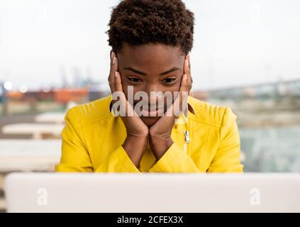 Visage surpris d'une femme afro-américaine noire en blouson jaune utilisant un ordinateur portable sur un bureau en bois dans la ville sur un arrière-plan flou Banque D'Images