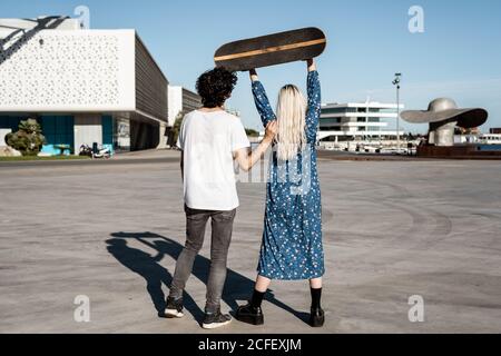 Vue arrière d'un jeune couple tendance méconnaissable debout tandis qu'une femme lève un skateboard sur une place contre un ciel bleu et des bâtiments modernes flous par temps venteux Banque D'Images