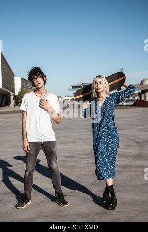 Jeune couple aimant tendance debout tandis que la femme tient un skateboard sur un carré contre le ciel bleu et des bâtiments modernes flous par temps venteux Banque D'Images