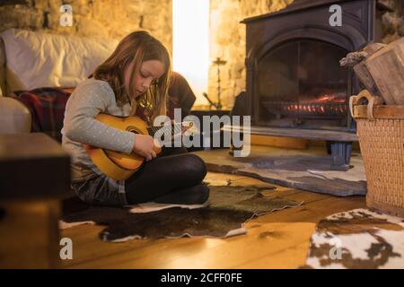 Jeune fille concentrée dans des vêtements décontractés assis avec des jambes croisées sur un canapé confortable et jouer ukulele dans la maison en pierre pendant Vacances en Cantabrie Banque D'Images