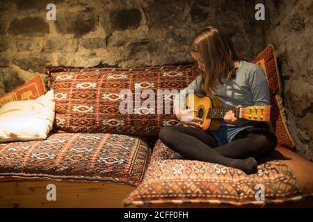 Jeune fille concentrée dans des vêtements décontractés assis avec des jambes croisées sur un canapé confortable et jouer ukulele dans la maison en pierre pendant Vacances en Cantabrie Banque D'Images