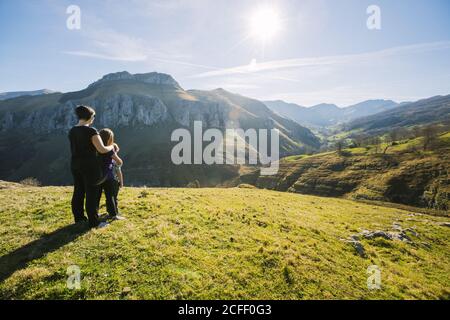 Vue arrière d'une randonneur anonyme qui embrasse les enfants en étant debout sur la prairie verte et l'observation pittoresque paysage de montagnes sur Journée ensoleillée en Cantabrie Banque D'Images