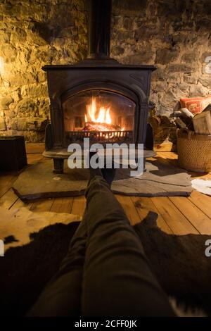 personne anonyme dans des vêtements décontractés assis avec les jambes tendues près cheminée dans le salon confortable de la vieille maison en pierre Banque D'Images