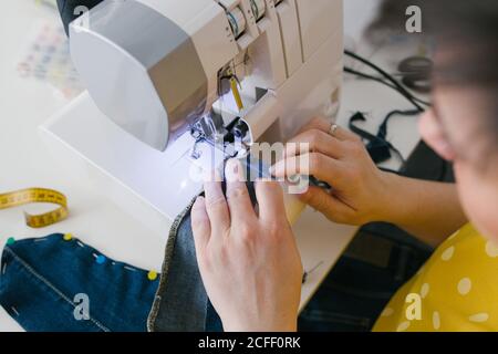 Par dessus, une femme brune non reconnaissable et coupée utilise une machine à coudre pour confecrer un vêtement en denim lors d'un atelier à domicile Banque D'Images