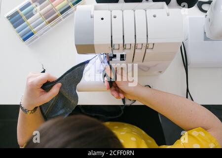 Par dessus, une femme brune non reconnaissable et coupée utilise une machine à coudre pour confecrer un vêtement en denim lors d'un atelier à domicile Banque D'Images