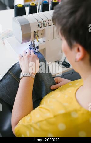 Par dessus, une femme brune non reconnaissable et coupée utilise une machine à coudre pour confecrer un vêtement en denim lors d'un atelier à domicile Banque D'Images