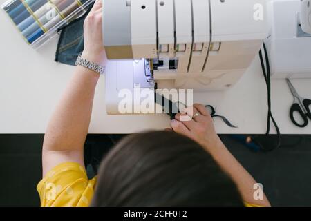 Par dessus, une femme brune non reconnaissable et coupée utilise une machine à coudre pour confecrer un vêtement en denim lors d'un atelier à domicile Banque D'Images