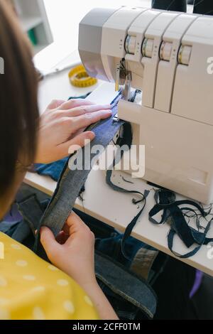 Par dessus, une femme brune non reconnaissable et coupée utilise une machine à coudre pour confecrer un vêtement en denim lors d'un atelier à domicile Banque D'Images