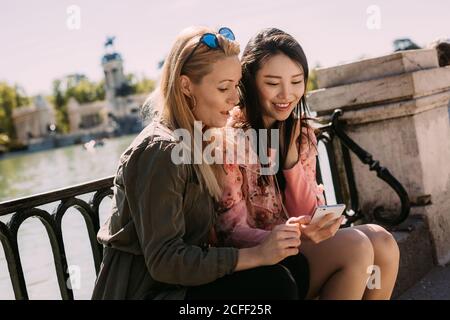 Jeunes femmes multiraciales dans des tenues à la mode souriant et parcourant un smartphone tout en étant assis près de la rampe de remblai le jour ensoleillé de la ville rue Banque D'Images
