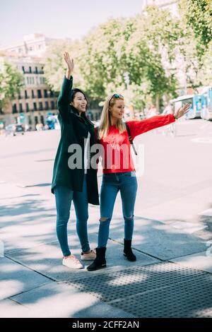 Joyeuses dames multiraciales dans des tenues élégantes souriant et en agitant les mains tout en étant debout sur la rue de la ville le jour ensoleillé ensemble Banque D'Images