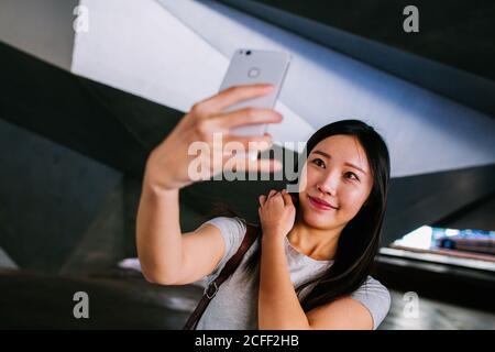 Jolie femme asiatique dans une tenue décontractée souriant et posant pour selfie dans un passage souterrain salopant Banque D'Images