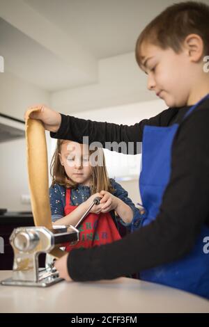 Une petite fille avec un frère plus âgé utilisant une machine à pâtes pendant préparation de nouilles maison dans la cuisine maison Banque D'Images