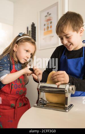 Une petite fille avec un frère plus âgé utilisant une machine à pâtes pendant préparation de nouilles maison dans la cuisine maison Banque D'Images