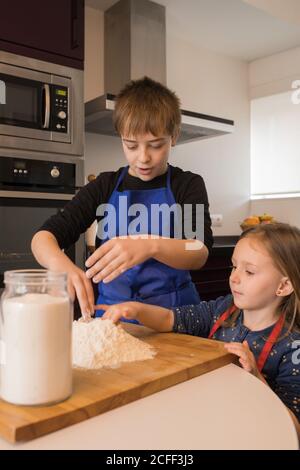 Une petite fille adorable aide un garçon plus âgé à préparer de la pâte pendant debout ensemble à la table de cuisine avec de la farine Banque D'Images