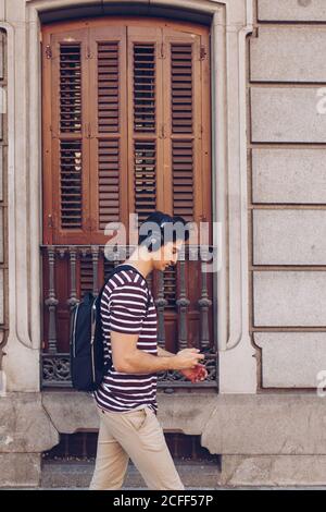Jeune homme attrayant dans des vêtements décontractés écoutant de la musique pendant la marche dans la rue en plein jour Banque D'Images