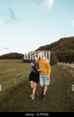 Joyeux couple romantique tenant les mains et marchant sur le vert champ en bord de mer au coucher du soleil avec ciel nuageux Banque D'Images