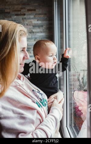 Jeune femme transportant un bébé mignon et regardant par la fenêtre le jour de pluie à la maison Banque D'Images